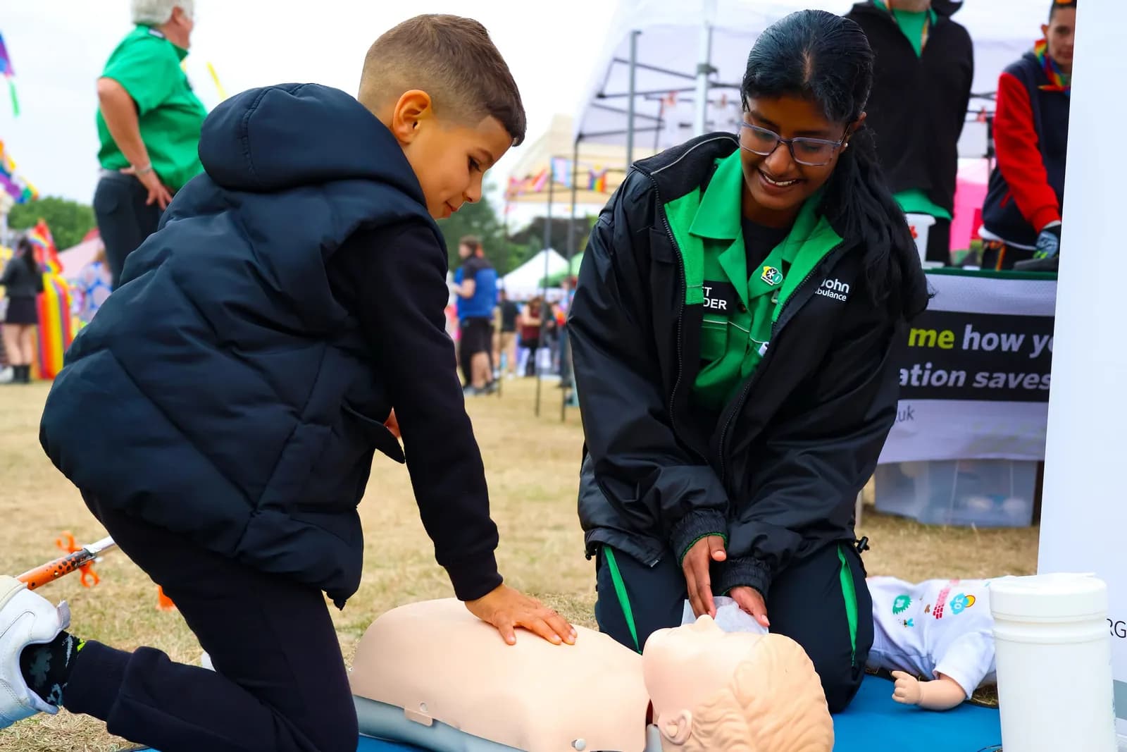 A smiling young woman in a green uniform teaches a boy how to perform CPR on a dummy.